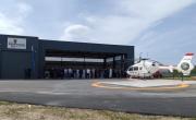 Un piste en bitume avec au centre un rond orange tracé puis un hélicoptère blanc et rouge. En arrière plan un hangar gris indiquant SDIS Pompiers de l'Allier et à ses portes une foule de gens dont certains en tenue d'infirmier