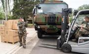 un militaire en treillis à droite conduisant un transpalette puis un militaire à gauche le guidant devant une palette de cartons. A l'arrière plan un camion de l'armée aux couleurs d'une tenue de camouflage