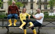 Un homme avec barbe blanche et lunettes à gauche, une jeune femme blonde aux cheveux blonds au centre et un jeune homme sont assis sur un banc dans la rue, avec des bottes jaunes de pluie aux pieds