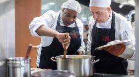 une femme avec une charlotte et un tablier de cuisine mélange quelque chose dans une casserole posée sur une table de cuisson, un homme avec une toque de chef et un tablier tient le couvercle de la casserole et observe