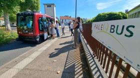 Des passagers montant dans le véhicule autonome, une petite navette rouge, dans une rue ensoleillée. le public prend des photos devant un panneau indiquant PaléoBus.