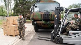 un militaire en treillis à droite conduisant un transpalette puis un militaire à gauche le guidant devant une palette de cartons. A l'arrière plan un camion de l'armée aux couleurs d'une tenue de camouflage