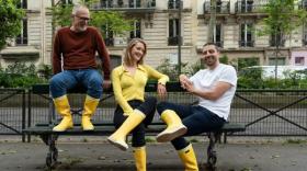 Un homme avec barbe blanche et lunettes à gauche, une jeune femme blonde aux cheveux blonds au centre et un jeune homme sont assis sur un banc dans la rue, avec des bottes jaunes de pluie aux pieds