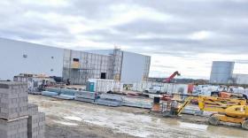 Une photo de chantier avec des matériaux (parpaings, câbles, poutres...) et des engins au premier plan. On voit un bâtiment industriel en construction sur la gauche et un bâtiment existant au fond à droite