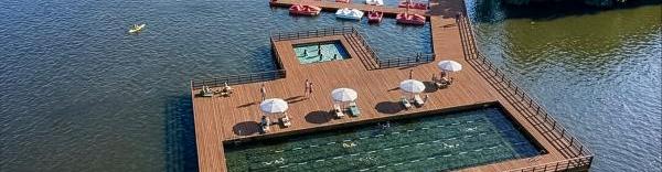 Une vue aérienne montrant la structure en bois au milieu de l'étang de Vieure. Au second plan une passerelle en bois ralliant la terre, avec un espace très vert. La plateforme en bois avec ses garde-corps,  entour un grand bassin avec des lignes de nage et un petit bassin où des enfants jouent. Des parasols et transat sont installés au bord des piscines et un ponton permet, à l'arrière plan d'amarrer des pédalos ou canoés. 