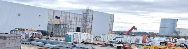 Une photo de chantier avec des matériaux (parpaings, câbles, poutres...) et des engins au premier plan. On voit un bâtiment industriel en construction sur la gauche et un bâtiment existant au fond à droite