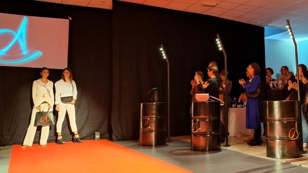 Un tapis rouge central accueille deux mannequins, dont miss Auvergne 2024 présentant chacune un sac cabas et demi-lune. un écran arbore le logo de la marque et à droite du public applaudit