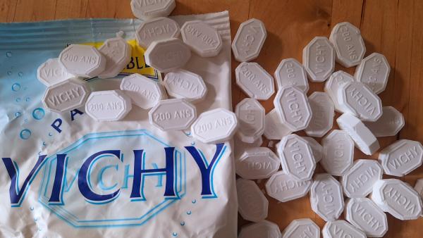 Un paquet de pastilles de Vichy ouvert sur une table en bois, des dizaines de pastilles blanches octogonales marquées Vichy ou 200 ans