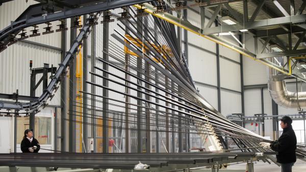 Deux femmes avec des casquettes et gants devant des barres d'aluminium montées en hauteur dans un hangar de travail