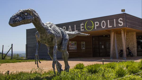 Une statue de dinosaure en grandeur réelle est installé dans l'herbe devant un bâtiment où l'on voit écrit Paléopolis sous un ciel bleu et du soleil