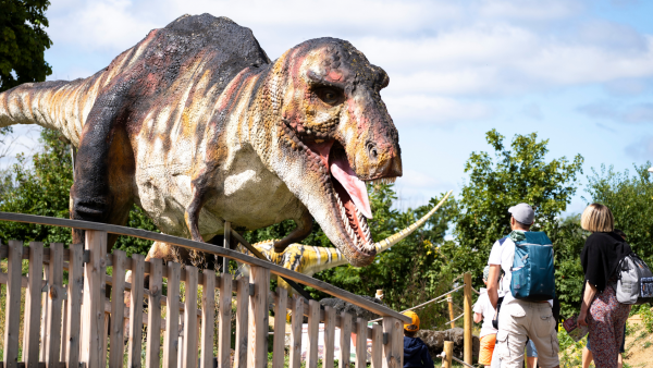 Dans un parc avec des arbres, un T-rex géant ouvre la gueule en direction d'une famille en balade, il y a deux enfants et un père et une mère avec sac à dos. Au second plan on aperçoit d'autres dinosaures en taille réelle.