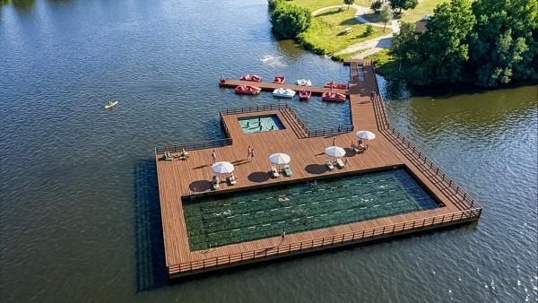 Une vue aérienne montrant la structure en bois au milieu de l'étang de Vieure. Au second plan une passerelle en bois ralliant la terre, avec un espace très vert. La plateforme en bois avec ses garde-corps,  entour un grand bassin avec des lignes de nage et un petit bassin où des enfants jouent. Des parasols et transat sont installés au bord des piscines et un ponton permet, à l'arrière plan d'amarrer des pédalos ou canoés. 