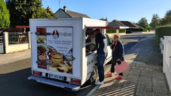 un camion de vente ambulante installé dans une rue. une femme âgée avec son cabas regarde les produits devant l'étal du camion, ouvert sur le côté. l'arrière du véhicule est floqué avec de illustrations de nourriture et le logo Patifrais