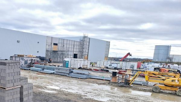 Une photo de chantier avec des matériaux (parpaings, câbles, poutres...) et des engins au premier plan. On voit un bâtiment industriel en construction sur la gauche et un bâtiment existant au fond à droite
