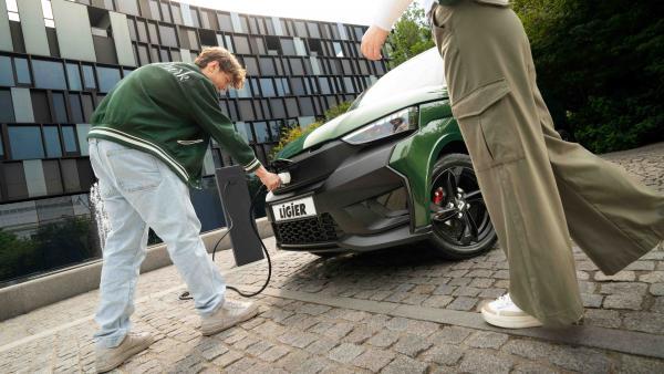 Un jeune homme penché sur sa voiture verte marqué Ligier sur sa plaque, est en train de recharger le véhicule depuis une borne de recharge. On distingue aussi les pieds d'une jeune femme à côté.